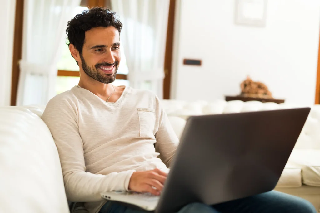 A smiling dark-haired man with a short beard sits casually on a white sofa looking down at a laptop on his lap.