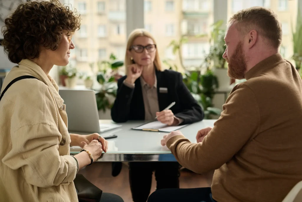 A casually dressed man and a woman in their 30s or 40s look at each other in front of a desk table. A woman in professional clothing looks at the man with her elbow leaning on the table and her chin resting on her closed fist. A laptop is open in front of her and she is holding a pen hovering over a clipboard.