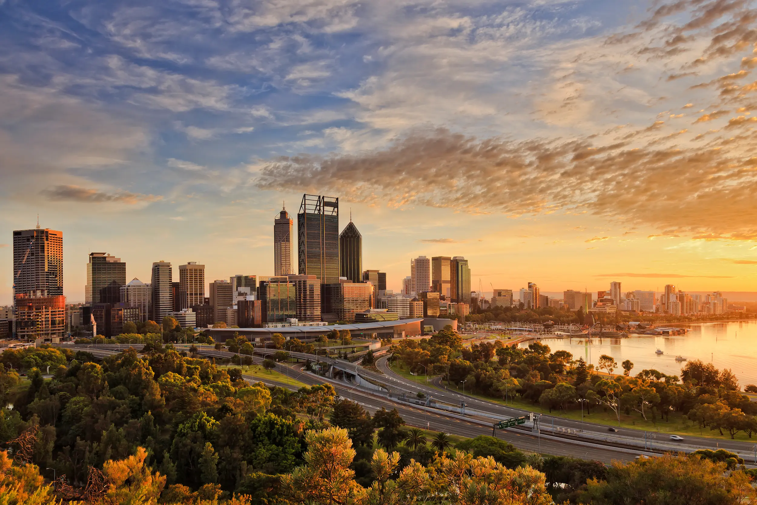 A view over Perth City from King's Park hill. The freeway in front of the city is visible, and the sun sets on right side of the photo, colouring the sky, clouds and the water in the Swan river a golden pink colour.