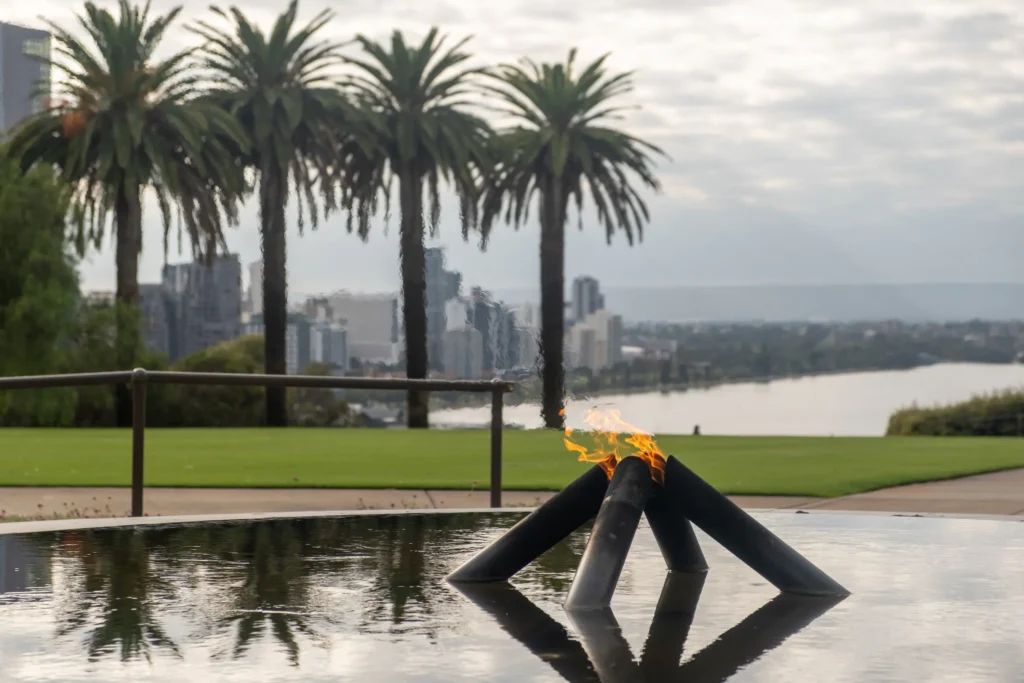 View of Perth City from King's Park, across a circular pond with a flame coming from four pipes emerging from the water. A strip of lawn and four palm trees separate the pond from the view over the city and the Swan River.