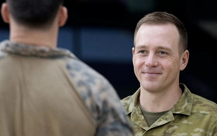 Head & shoulders view of a man with short dark hair in a camouflage shirt smiling with closed lips at a man with his back to the camera.