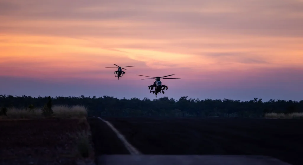 A sunset scene in which two army helicopters hover low over a darkened ground with pink colouring the clouds behind.
