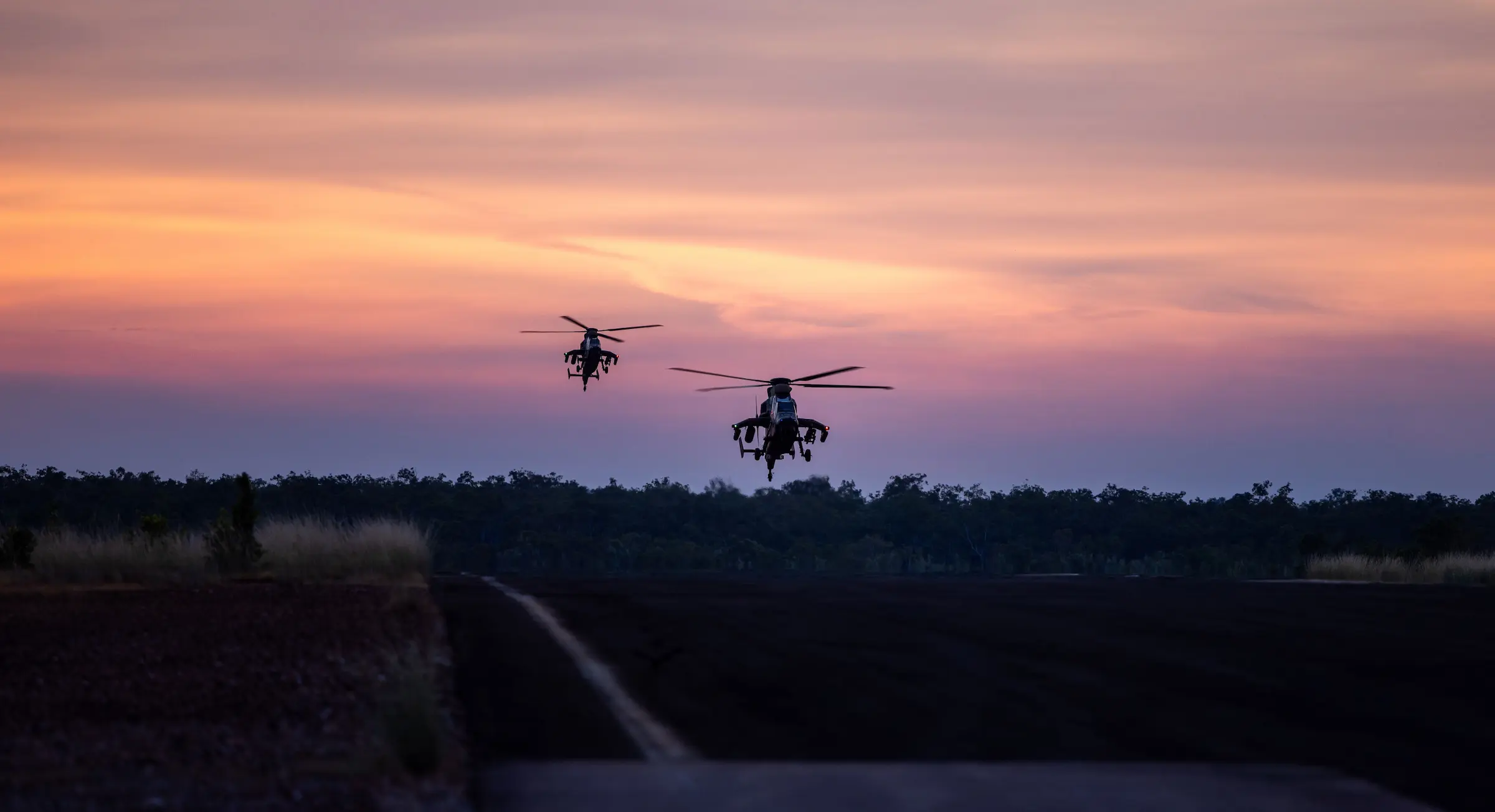 A sunset scene in which two army helicopters hover low over a darkened ground with pink colouring the clouds behind.