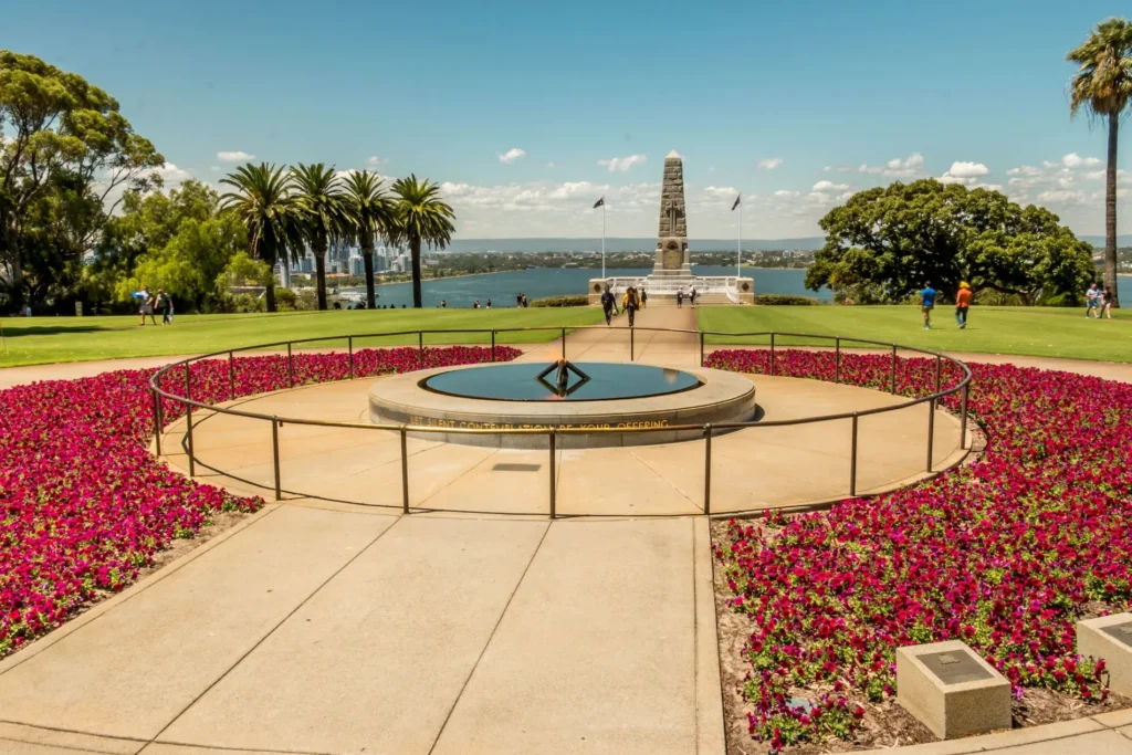 A view of the state war memorial in King's Park, Perth. Perth City and the Swan River is in the far background. In the foreground is a wide beige-coloured concrete path that goes toward and circles around both sides of the pond with a burning flame on pipes in the middle. The path then continues on into the distance toward an obelisk with flags on either side of it. Around the pond and path is mass-planted purple-red flowers that appear to be petunias. A light green lawn separates the pond area from the obelisk, and several people are walking on the lawn and on the path in the distance.