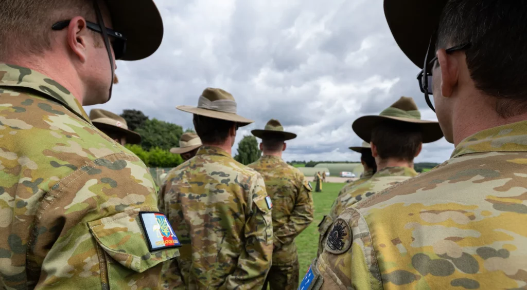 View from behind of several soldiers in camouflage uniforms and broad-brimmed hats standing at ease on a lawn with low clouds in the sky.