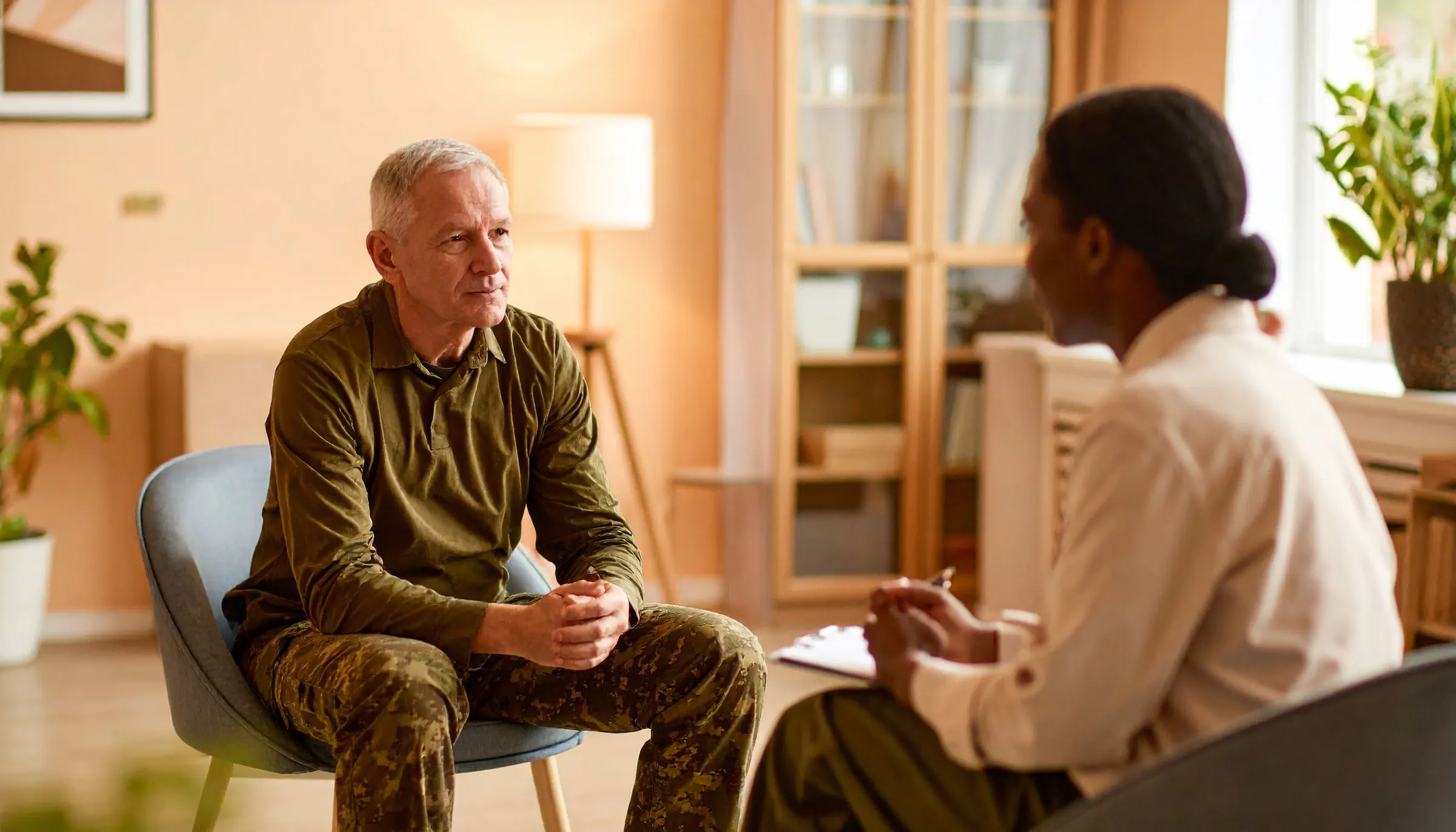 An older grey-haired man with military bearing in an olive green long-sleeved collared Henley shirt and camouflage trousers sits on a chair facing a dark-skinned woman with her back/side to the camera. The woman has her hair pulled back in a low bun and is wearing a cream-coloured shirt.