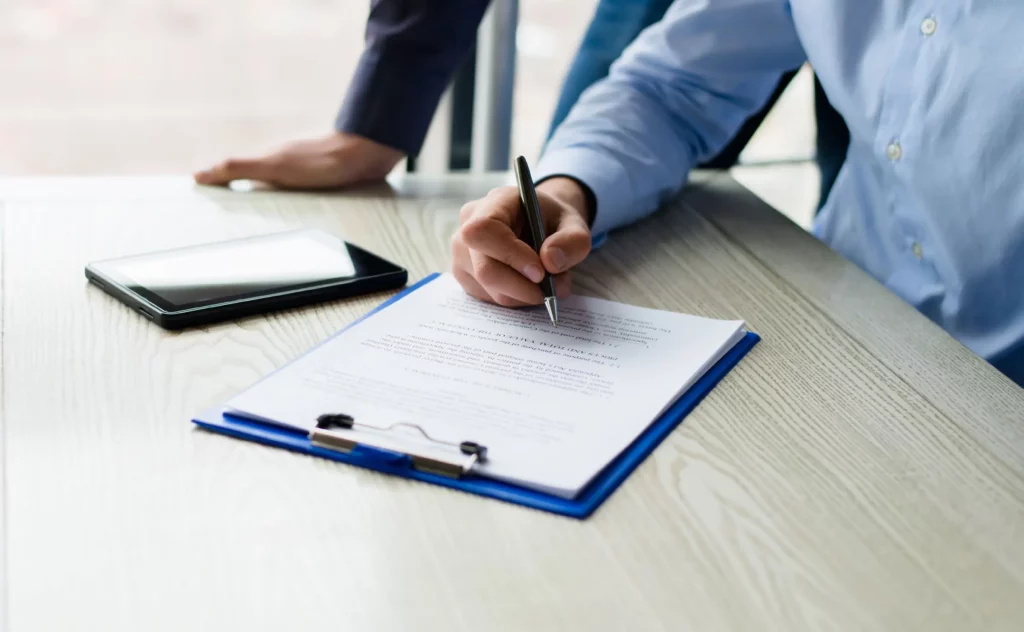 Close-up view of a man's hand and arm with the front of his shirt showing to the right of the picture. He is holding a pen over a paper on a clipboard. A large phone-like device lies next the the clipboard, and another man's hand is visible leaning on the table behind the first man.