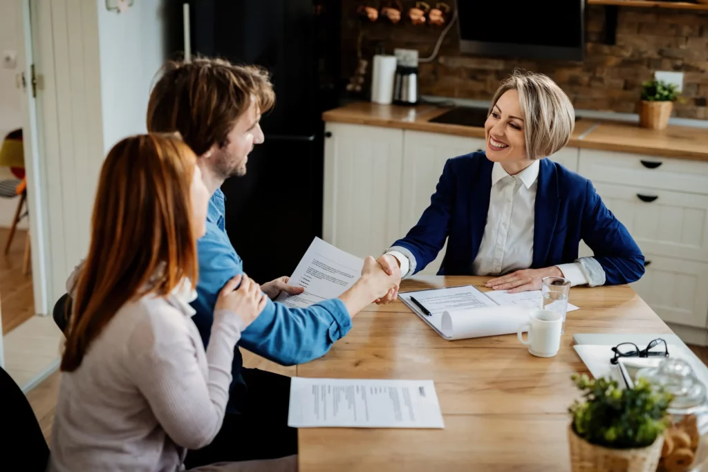 A cheerful-looking young couple sit at a table with a smiling businesswoman. The man holds a paper and shakes the hand of the businesswoman while the woman at his side places her hand on his arm. Paperwork and a cup and water glass are on the table, and behind is a cabinet with a sink and cupboards and drawers.