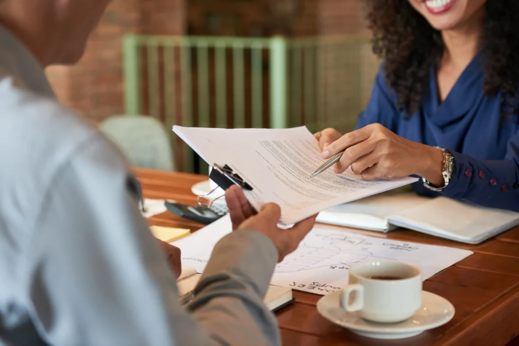 A partial view of a man and woman on either side of a desk. The man is grasping a sheaf of papers with a clip holding them together that the woman is holding from across the desk. The woman is holding a pen. On the desk are papers, a book, and a cup and saucer.