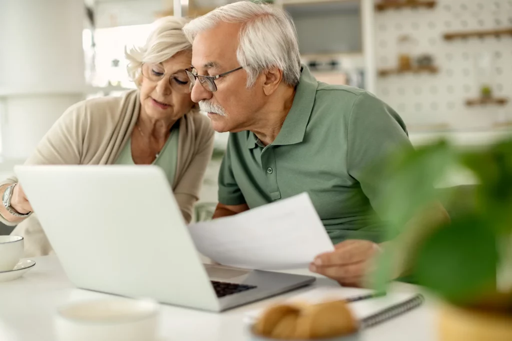 A seated senior couple lean with their heads together over a table with a laptop and paperwork. Cups and saucers sit on the table, along with a spiral-bound notebook and pen.