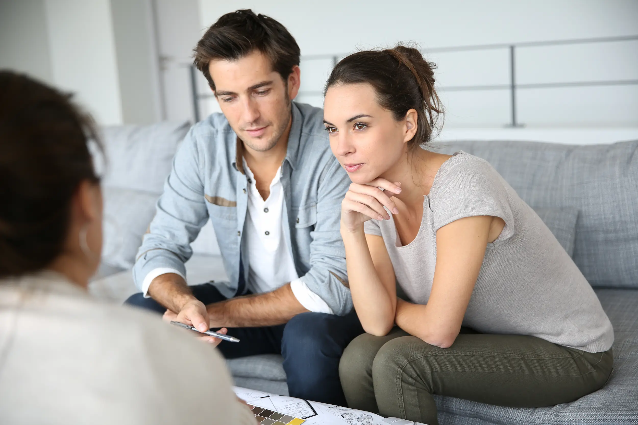 A young couple sit close together on a light-grey couch, facing a woman with her back to the camera. The woman has one elbow on her knee and has her chin resting on her hand. The man is holding a pen and looking down at something on a low coffee table in front of the couple.