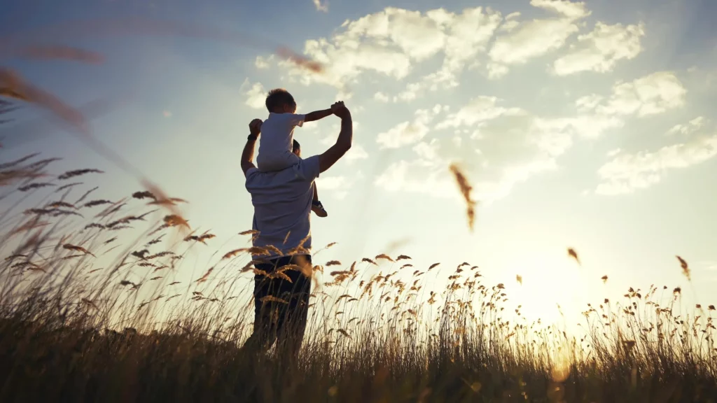 The back view of a man standing in a field holding the hands of a young child sitting on his shoulders. Stalks of grain line the foreground and a patch of clouds is in the sky above them. The bright light of a setting sun comes from the bottom right of the photo.